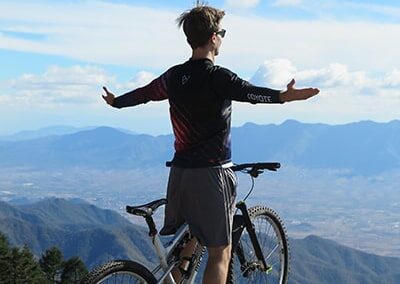 A man stands on a bicycle with his arms outstretched in front of a mountain view